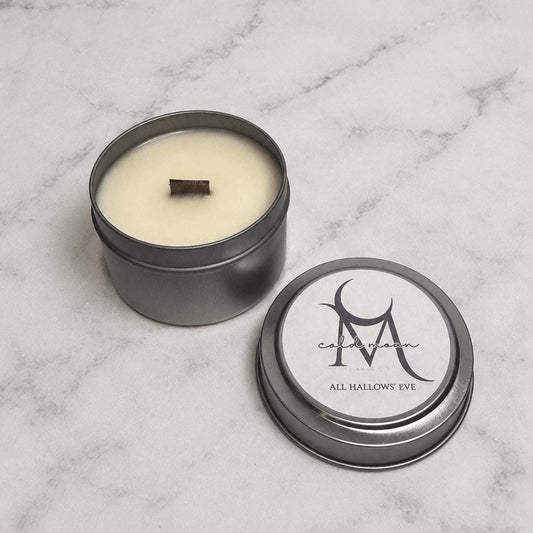 Open All Hallows’ Eve wood-wick tin candle beside its labeled silver lid on marble.