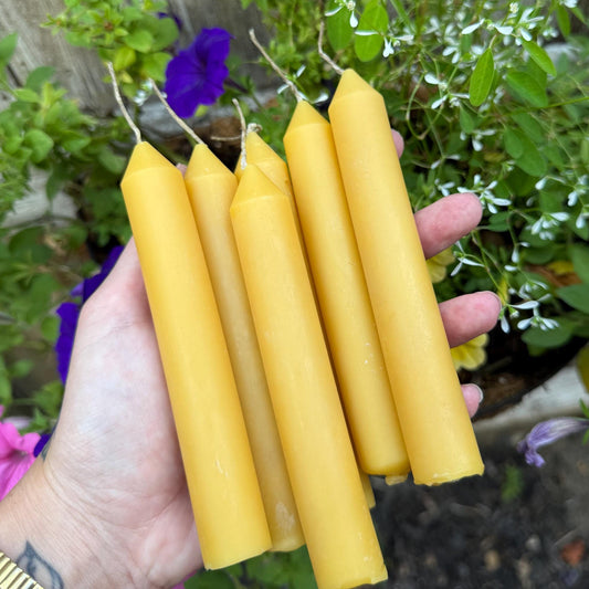 Golden beeswax taper candles held in a hand in front of garden flowers.