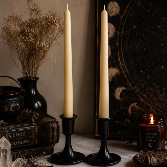 Two ivory soy taper candles in black holders on a ritual table with flowers and a lit jar candle.