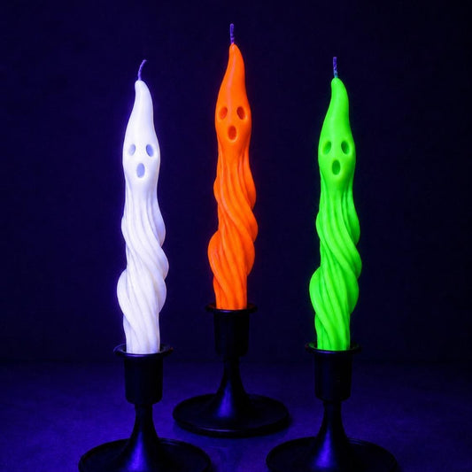 May include: Three taper candles shaped like ghosts, each featuring a twisted body and a carved face with two eyes and an open mouth. The candles are white, orange, and neon green, and each is held upright in a black metal candle holder base.